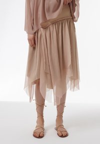 Falda asimétrica beige fluida con un diseño en capas y cinturilla fruncida, combinada con sandalias planas marrones con tiras trenzadas.