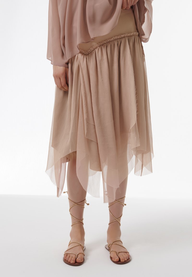 Falda asimétrica beige fluida con un diseño en capas y cinturilla fruncida, combinada con sandalias planas marrones con tiras trenzadas.