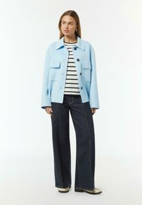 Giacca blu chiaro in misto lana con colletto a punta, tasche frontali e bottoni neri. Abbinata a una camicia a righe e pantaloni in denim scuro.