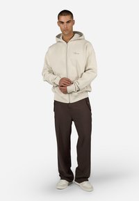 Lys beige zip-up hettegenser med hette, med en frontlomme. Kombinert med mørkebrune, løstsittende bukser og hvite joggesko. Glatt tekstur.
