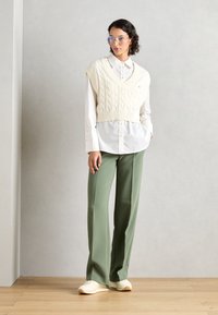 Krämfärgad kabelstickad ärmlös tröja över en vit skjorta, matchad med gröna vida byxor och ljusa sneakers. Klara glasögon bärs.