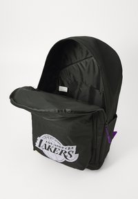 Mochila preta com fecho de zíper, apresenta um grande gráfico branco que diz "LAKERS" e um puxador de zíper roxo. O interior é forrado com tecido preto.