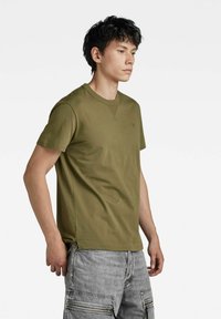 T-shirt a girocollo verde oliva realizzato in cotone, con maniche corte e un logo discreto sul petto. Indossato con pantaloni cargo grigi.