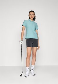 Světle modrá polo tričko s krátkým rukávem a límečkem, nošené s tmavými šortkami. V levé ruce drží bílé boty a golfovou holi.