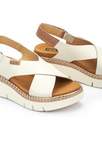 Sandalias de cuero blanco con correas cruzadas, plantilla acolchada beige, suela de plataforma de goma texturizada y un acento de cuero marrón en el talón.