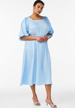 Kvinde iført en lyseblå midi kjole med sorte polkaprikker og puffede ærmer, stående barfodet mod en hvid baggrund.