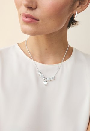 Frau trägt eine silberne Charms-Halskette mit Perlen und Herzanhänger, kombiniert mit passenden silbernen Tropfenohrringen und einer weißen Bluse.