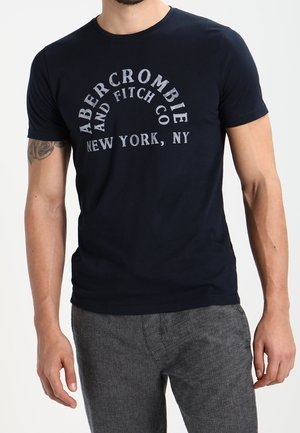 Homme portant un t-shirt bleu marine Abercrombie & Fitch New York, NY et un pantalon gris, montrant un tatouage sur son bras supérieur gauche.