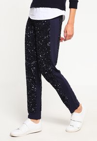 Marinblå glittriga träningsbyxor har en slim fit med slätt tyg på sidorna och glittrande dekor, kombinerat med vita sportskor.