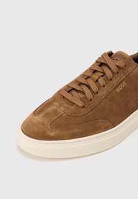 Καφέ σουέντ αθλητικό παπούτσι με στρογγυλερή μύτη και λευκή ελασ τική σόλα. Διαθέτει κορδόνια στο ίδιο χρώμα και ανάγλυφο λογότυπο στο πλάι. Απλός σχεδιασμός.