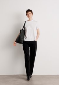 White cotton t-shirt with embroidered light blue logo, paired with black trousers and a black leather tote bag. Smooth textures, casual fit.