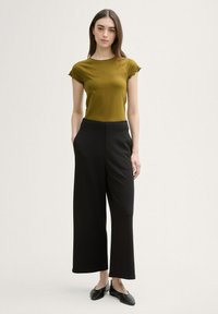 Top verde măsliniu cu mâneci scurte și margini fâlfâitoare, asortat cu pantaloni negri din material lejer, cu talie înaltă. Modelul poartă, de asemenea, pantofi negri fără toc.