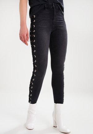Persoon die zwarte skinny jeans draagt met veters aan de zijkanten en witte enkellaarsjes met hakken, staand tegen een effen lichte achtergrond.