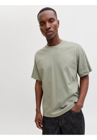 Νέος άνδρας με κοντά μαλλιά, ντυμένος με πράσινο χλωρό κοντομάνικο μπλουζάκι και σκούρο τζιν, στέκεται με το ένα χέρι στην τσέπη μπροστά σε απλό φόντο.