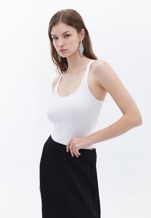 Débardeur blanc côtelé avec bretelles larges, associé à une jupe noire. Les boucles d'oreilles du modèle sont grandes, angulaires et en métal argenté.