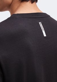 Gros plan d'une personne portant un t-shirt noir avec un logo Reebok blanc vertical dans le haut du dos, près de l'encolure.