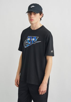 Young man wearing a black Nike Air t-shirt with blue graphic and black Nike cap, standing against a plain light gray background.