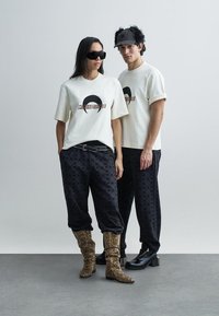 White t-shirts with graphic design, paired with black patterned pants. Model wears sunglasses and snake-print boots. Background is minimal.