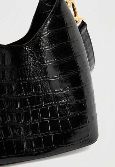 Μαύρη δερμάτινη τσάντα χειρός με υφή κροκόδειλου και χρυσό μεταλλικό κούμπωμα, εμφανίζεται σε κοντινή λεπτομέρεια πάνω σε λευκό φόντο.