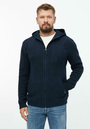 Mann mit Bart, der einen marineblauen Kapuzenpullover mit Reißverschluss und hellblaue Jeans trägt, steht mit einer Hand in der Tasche vor weißem Hintergrund.
