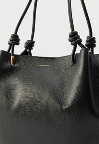 Borsa tote in pelle nera con finitura testurizzata, caratterizzata da dettagli con nodi sulle maniglie e un accento logo in metallo dorato. Forma semplice e strutturata.