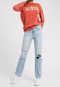 Rød langærmet skjorte med "GUESS" i hvid tekst, parret med lyseblå slidte jeans, som har hul nær begge knæ, og hvide sneakers.