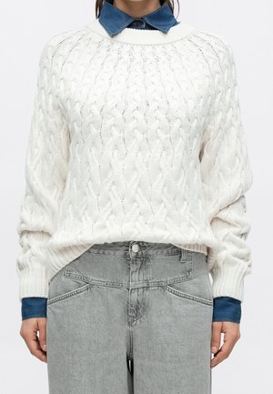 Persona con un jersey blanco de punto trenzado sobre una camisa azul con cuello y vaqueros gris claro de talle alto, de pie contra un fondo liso.