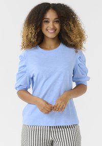 Kék, rövid ujjú felső, kerek nyakkivágással és puffos ujjal; fekete-fehér csíkos nadrággal viselve, sima szövetből készült.