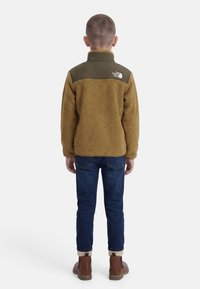Enfant de dos portant une veste polaire vert olive, un jean bleu foncé, des chaussettes beiges et des bottes marron sur fond blanc.