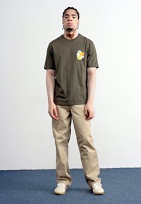 Oliven grøn bomuld t-shirt med et flerfarvet grafisk motiv på venstre bryst, parret med beige bukser og hvide sko. Enkel, afslappet design.