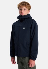 Marineblaue Zip-Jacke mit Kapuze, featuring elastische Bündchen, einem Logo-Patch auf der Brust und einem leichten Material. Einfaches Design und Schnitt.