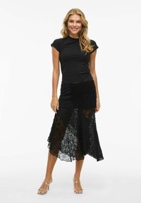 T-shirt noir ajusté associé à une jupe en dentelle noire longueur sol avec un design superposé et des accents texturés. Sandales claires et élégantes.