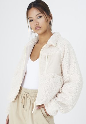 Veste en polaire beige avec un tissu doux et texturé, fermeture éclair à l'avant et un panneau lisse sur la poitrine. Présente un col haut et des poches latérales.