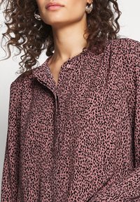 Blouse rose boutonnée avec imprimé tacheté noir, dotée d'un col et d'une texture de tissu douce. Cheveux bouclés et grandes boucles d'oreilles visibles.