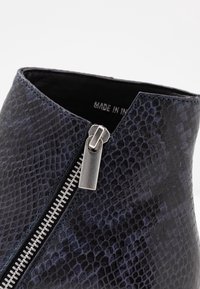 Stivaletto realizzato in materiale testurizzato con stampa pitone nera, dotato di una cerniera laterale metallica e cuciture blu in contrasto. Etichetta interna visibile.