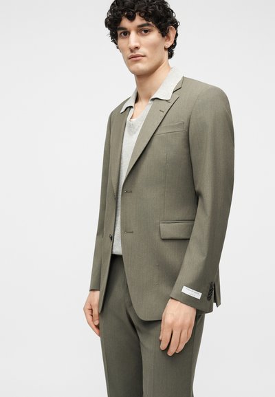 Άνδρας με σγουρά μαλλιά που φοράει ελιά πράσινο κοστούμι και ανοιχτό γκρι πουλόβερ, στέκεται μπροστά σε απλό φωτεινό φόντο.