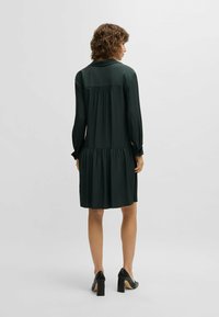 Dunkelgrünes Kleid aus glattem Stoff, mit langen Ärmeln, einem gefältelten Rückendesign und einem ausgestellten Saum. Getragen mit schwarzen Absatzschuhen.