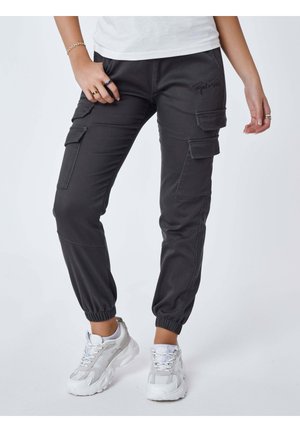 Person iført sorte cargobukser med elastiske manchetter og hvide sneakers, stående med den ene hånd i lommen og den anden afslappet ved siden.