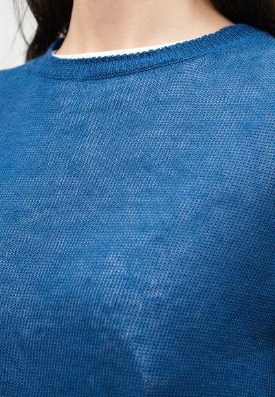 Gros plan d'une personne portant un pull tricoté bleu texturé avec un col rond et des cheveux foncés visibles au niveau du col.