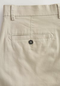 Beige Hose mit glatter Textur, ausgestattet mit einer Gesäßtasche mit einem schwarzen Knopf und Gürtelschlaufen am Bund.