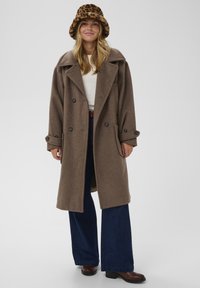 Hnedý kabát z vlnenej zmesi s dvojradovými gombíkmi, veľkým golierom a bočnými vreckami, doplnený tmavomodrými rozšírenými džínsovými nohavicami a klobúkom typu bucket s leopardím vzorom.