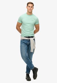 T-shirt in cotone verde con maniche corte, abbinato a jeans in denim blu. Maglione grigio legato in vita e stivali neri con lacci.