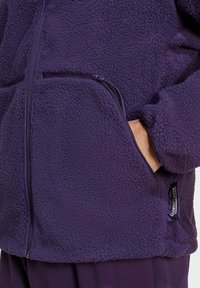Giacca in pile viola con superficie texurizzata, dotata di zip frontale e tasche laterali. Etichetta di branding evidente sulla cucitura inferiore.