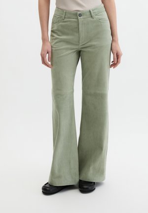 Gros plan sur les jambes d'une personne portant un pantalon large vert clair et des chaussures noires, debout sur un fond blanc uni.