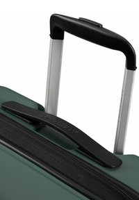 Valigia rigida di colore verde scuro con finitura texture, dotata di zip nera, maniglia nera e manico telescopico argentato.