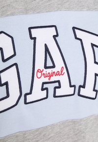 Grå och ljusblå bomullstyg med stora vita bokstäver som stavar "GAP" med röd sömnad på ordet "Original" nedan.