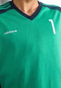 Vihreä Adidas-urheilupaita, jossa on tummansininen V-kaula-aukko, kolme tummansinistraipua olkapäällä ja valkoinen numero "1" oikeassa rinnassa.