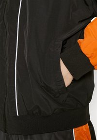 Black bomber jacket with an orange sleeve, featuring a white zip closure, ribbed cuffs, and a smooth, glossy texture.