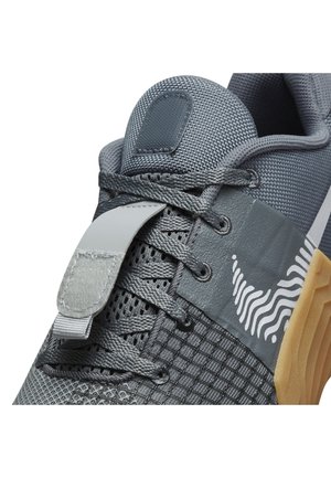 Gros plan d'une chaussure de sport grise avec textures en mesh et tissu, logo ondulé blanc sur le côté, semelle en caoutchouc beige, montrant les lacets et la languette rembourrée.