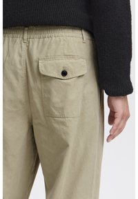 Pantalon beige clair avec une ceinture élastique, une poche sur la couture latérale et une poche arrière boutonnée. Le tissu semble doux et texturé.